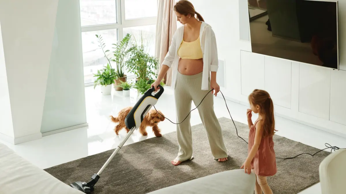 Mulher aspirando o tapete da sala enquanto a filha brinca com o cachorro