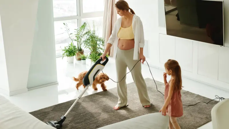 Mulher aspirando o tapete da sala enquanto a filha brinca com o cachorro